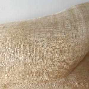 May include: A close-up shot of a tan woven fabric. The material has a textured surface and a loose weave, giving it a slightly transparent look. The fabric appears to be a natural fibre, possibly linen.