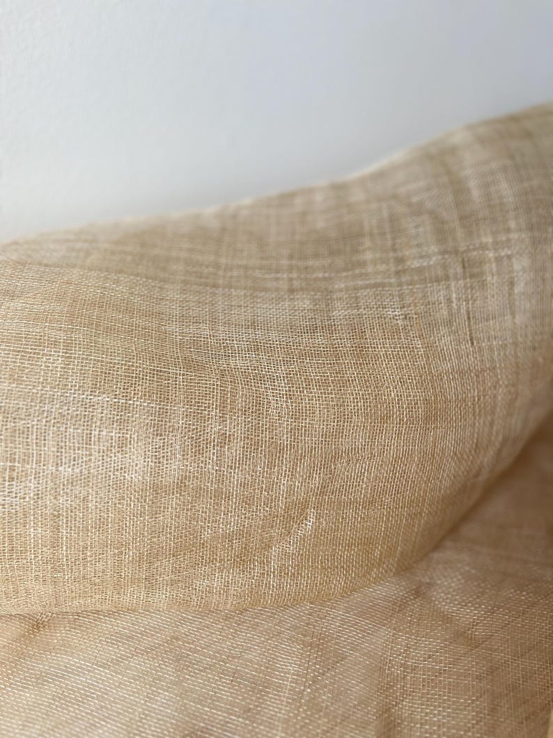 May include: A close-up shot of a tan woven fabric. The material has a textured surface and a loose weave, giving it a slightly transparent look. The fabric appears to be a natural fibre, possibly linen.