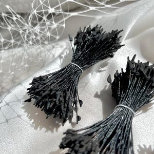 May include: Two bundles of black plastic price tags, secured with white wire ties. The tags are on a white satin surface, with a white net-like fabric in the background. Each tag has a small, teardrop-shaped end.