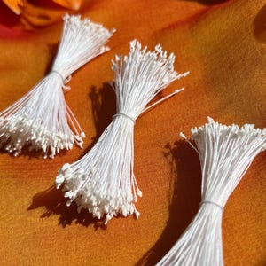 May include: Three bundles of white stamens, tied with thin string, are arranged on orange and yellow fabric. Each bundle has numerous thin, white stems with small, white, bead-like tips. The background is a gradient of orange and yellow.