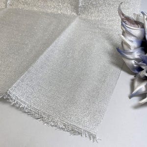 May include: A close-up of a shimmering silver fabric with a fringed edge. The fabric is folded, showing its texture and sheen. A decorative flower with blue and white petals is in the background.