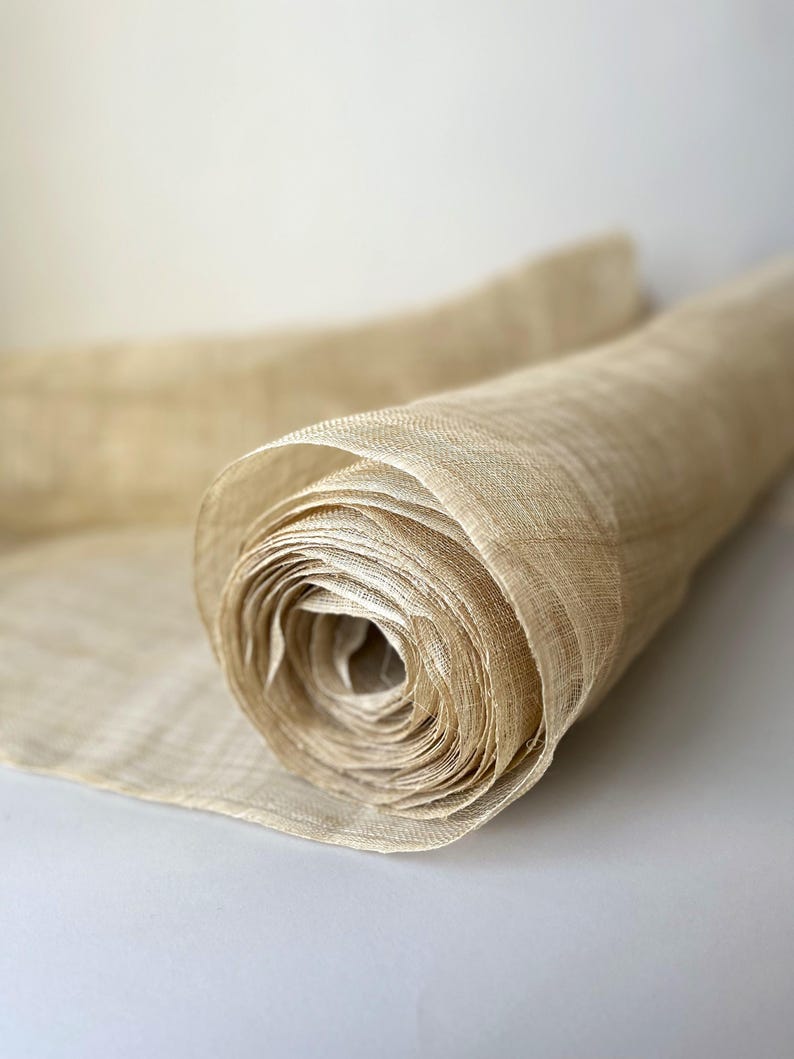 May include: A rolled-up length of beige fabric. The material has a woven texture and looks to be made from natural fibres. The roll is partially unrolled, showing the inner layers. The background is a plain, light colour.