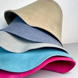 May include: A stack of felt fabric in various colors, including tan, blue, gray, turquoise, and fuchsia. The felt is layered, showcasing the different hues and textures. The image is well-lit, highlighting the soft, smooth surfaces of the felt.
