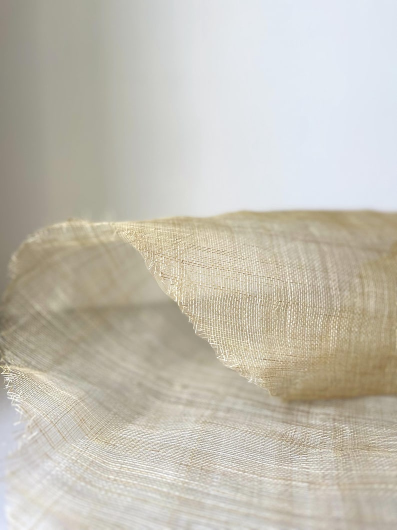 May include: Close-up of a section of woven fabric in a light beige colour. The fabric has a delicate, open weave, with visible threads creating a textured surface. The edges of the fabric are slightly frayed, adding to its natural appearance.