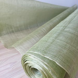 May include: A roll of pale green fabric is shown, possibly linen or a similar material. The fabric is tightly rolled, revealing layers of the material. The texture appears slightly coarse, with visible woven threads. The colour is a soft, muted green.