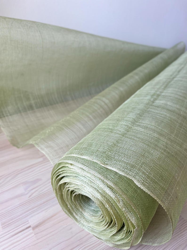 May include: A roll of pale green fabric is shown, possibly linen or a similar material. The fabric is tightly rolled, revealing layers of the material. The texture appears slightly coarse, with visible woven threads. The colour is a soft, muted green.
