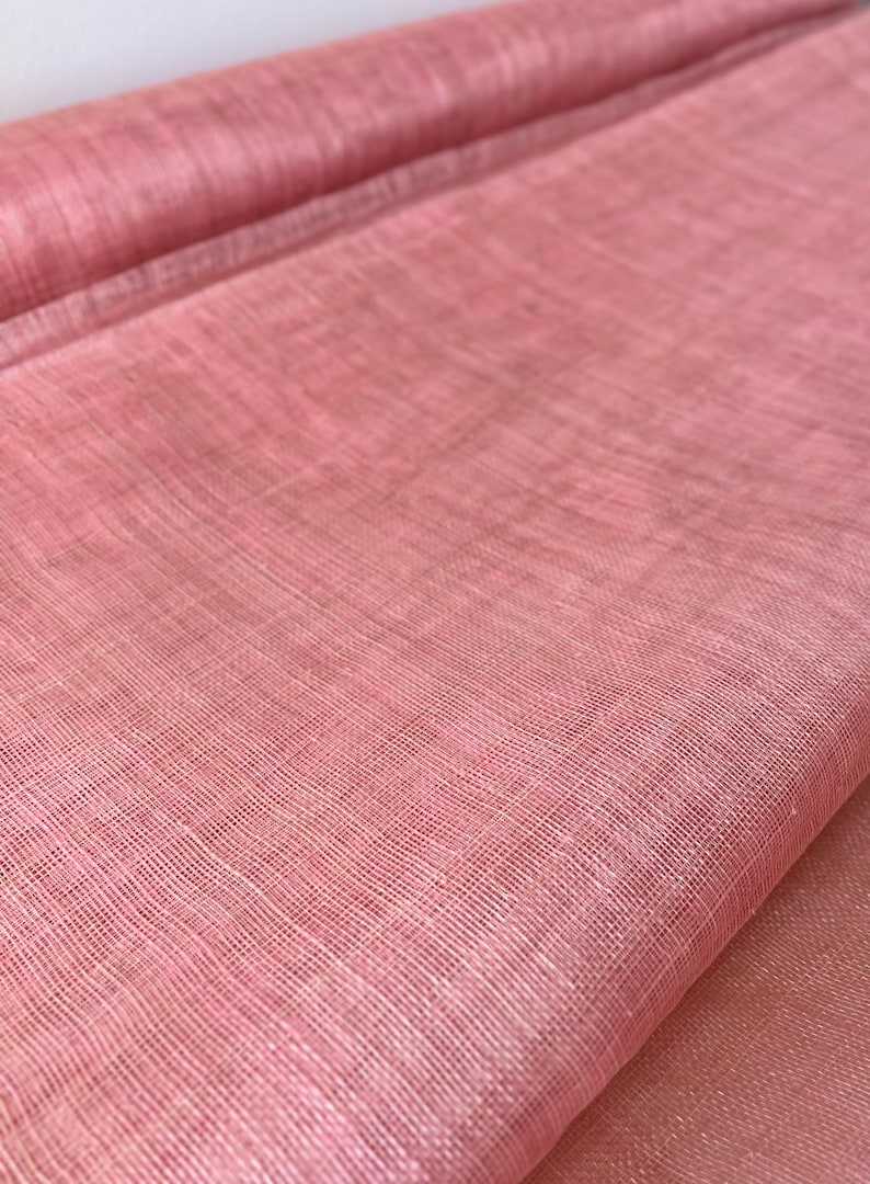 May include: A roll of pink fabric with a woven texture. The fabric is a light, dusty rose colour and appears to be lightweight. The weave is visible, creating a grid-like pattern. The fabric is likely used for clothing or home decor.
