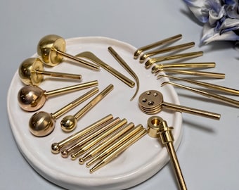 May include: A collection of gold-toned metal sculpting tools arranged on a white ceramic tray. The set includes various ball styluses, curved picks, a multi-hole disc tool, and a cylindrical punch, all featuring smooth, polished metallic surfaces.