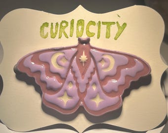 May include: A decorative plaque featuring a stylized moth design in shades of pink and lavender, with yellow star and crescent moon accents. The word "CURIOCITY" is written in green above the moth. The plaque has a cream-colored background.