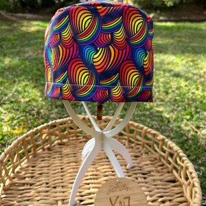 May include: A patterned cap with a rainbow design of curved lines. The cap is displayed on a white stand, set on a woven tray. A wooden tag with the text "XNZ Handmade with Love" hangs from the stand.