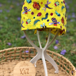 May include: A yellow scrub cap featuring a vibrant insect design, including bees, butterflies, and dragonflies. The cap is displayed on a white stand, with a wooden tag that reads "XNZ Handmade with Love".