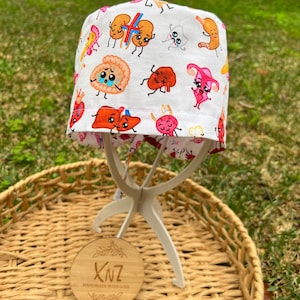 May include: A white surgical scrub cap with colourful cartoon illustrations of human organs. The cap is displayed on a white stand, set against a backdrop of green grass and a woven basket. The cap has a red trim.