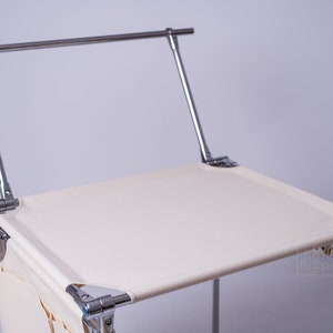 May include: A portable makeup station featuring a cream-coloured fabric seat and a chrome metal frame. The structure includes a horizontal bar at the top and a side pocket made of the same cream fabric. The text Photo Props is visible on the seat.