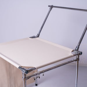 Puede incluir: Un soporte de telón de fondo blanco con un marco cromado y una barra horizontal. El soporte está diseñado para la fotografía y tiene una bolsa de tela unida a un lado.