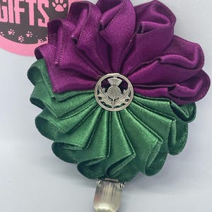 Könnte beinhalten: Handgefertigtes, blumenförmiges Accessoire in Lila und Grün mit einem silbernen Distelemblem in der Mitte. Das Accessoire hat eine Metallklammer auf der Rückseite. Es handelt sich wahrscheinlich um eine Brosche oder einen dekorativen Clip.