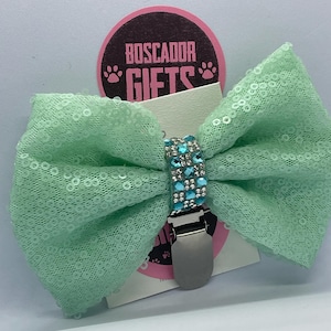 Puede incluir: Un lazo de lentejuelas verde menta con un clip plateado y un centro de gemas azules brillantes. El lazo está sujeto a una etiqueta con el texto "BOSCADOR GIFTS" en un círculo rosa.