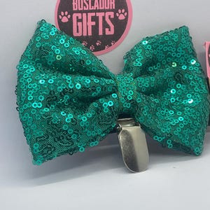 Può includere: Un fiocco verde brillante con paillettes e una clip argentata. Il fiocco è davanti a un logo circolare rosa e nero che dice "Boscador Gifts". Il fiocco è probabilmente per un animale domestico o un bambino.