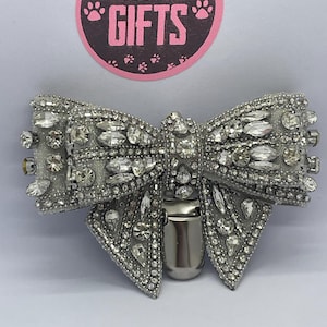 May include: A silver-toned, bow-shaped pet collar charm, lavishly decorated with clear rhinestones. The charm features a metal clip at the back and is set against a white backdrop. The text "BOSCADOR GIFTS" is displayed in a pink circle.