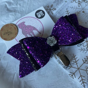 May include: Purple glitter bow with a silver flower accent and a silver clip. The bow is on a white background with snowflakes.