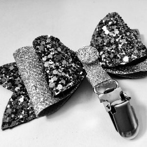 May include: A black and silver glitter bow with a silver clip. The bow is made of layered glitter fabric and has a round, silver embellishment in the centre. The clip is silver and attached to the bow.