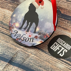 Peut inclure: Une décoration ronde et plate représentant la silhouette noire d'un chien portant un bonnet de Père Noël rouge et blanc. Le fond montre une scène enneigée avec des branches de pin et des baies rouges. Le nom Nelson est écrit en noir.
