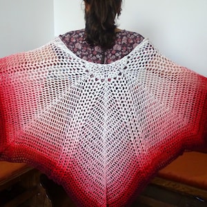 Pode incluir: Um poncho de crochê branco com um design degradê vermelho e branco. O poncho é feito com um ponto solto e aberto e tem uma forma ampla e fluida.
