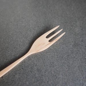 Może przedstawiać: Jasny drewniany widelec z trzema zębami. Widelec jest ustawiony pod kątem ukośnym na szarej powierzchni. Widoczne jest usłojenie drewna, a widelec wydaje się być wykonany ręcznie. Tło jest jednolite, neutralne.