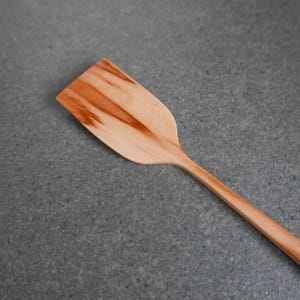 Könnte beinhalten: Ein Holzspatel mit rechteckigem Kopf und langem, schlankem Griff. Der Spatel besteht aus hellem Holz mit dunkleren Maserungsmustern. Der Spatel ist diagonal über einer grauen Oberfläche angewinkelt.