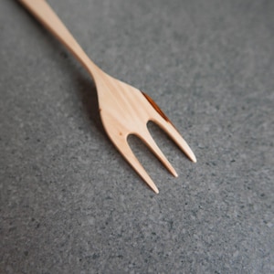 Może przedstawiać: Drewniany widelec z trzema zębami, wykonany z jasnego drewna. Uchwyt rozciąga się w lewo, a widelec spoczywa na szarej powierzchni. Widoczne jest usłojenie drewna.