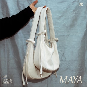 May include: Off-white shoulder bag with a curved design, featuring a zipper closure and long straps. The bag is held by a person wearing a black sleeve. The word "MAYA" is printed in a serif font. The text "pdf sewing pattern" is also visible.