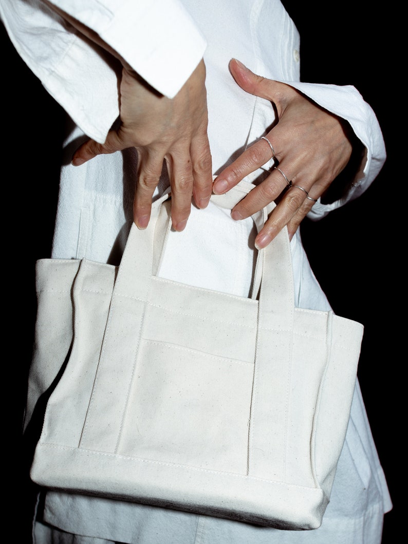 May include: A small, off-white canvas tote bag featuring two sturdy top handles, a rectangular base, and a flat front pocket with visible vertical stitching details. The bag is constructed from a plain, light-colored fabric with a structured shape.