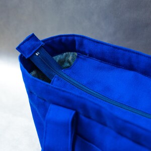 May include: A vibrant royal blue canvas tote bag featuring a secure top zipper closure. The bag has sturdy fabric handles and a visible interior lining with a subtle grid pattern, providing a clean and functional design for daily use.