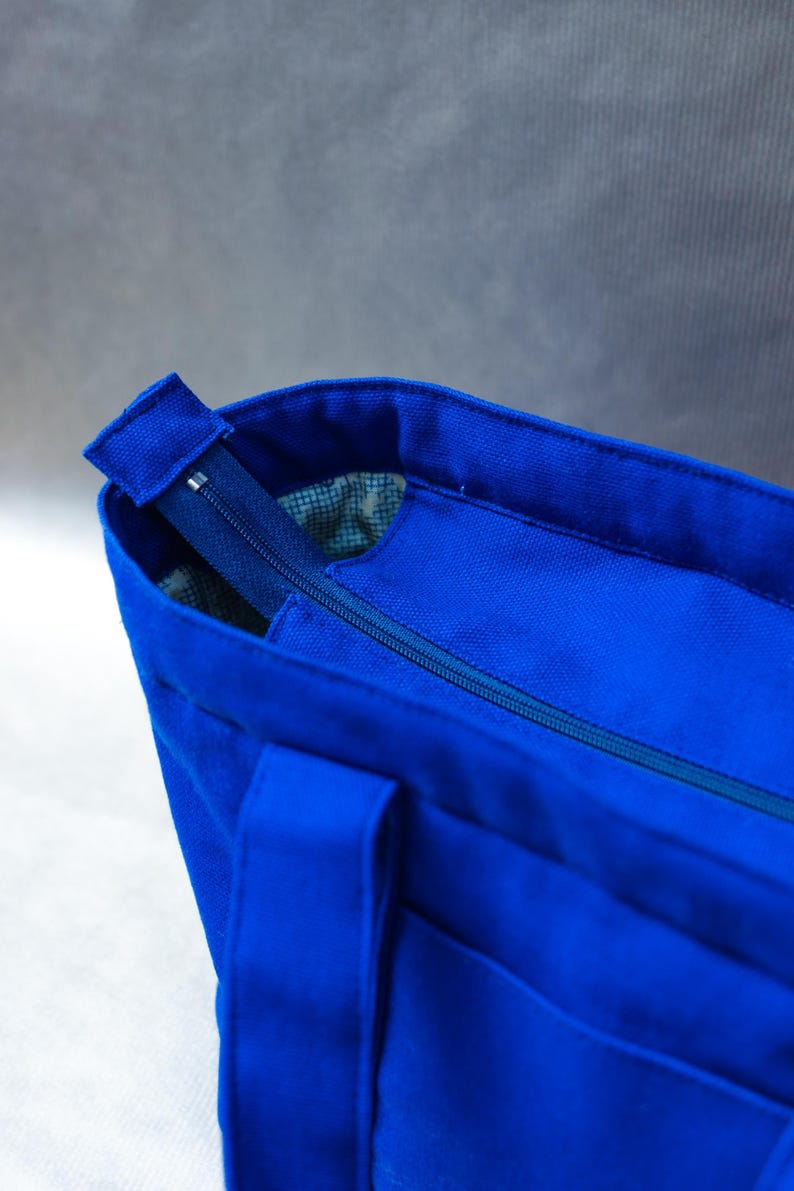 May include: A vibrant royal blue canvas tote bag featuring a secure top zipper closure. The bag has sturdy fabric handles and a visible interior lining with a subtle grid pattern, providing a clean and functional design for daily use.