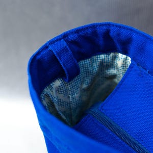 May include: A bright blue canvas tote bag featuring a structured opening with a matching blue zipper closure. The interior is lined with a white fabric that has a small blue grid pattern, and a blue fabric loop is attached to the rim.