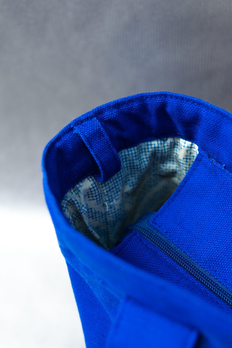 May include: A bright blue canvas tote bag featuring a structured opening with a matching blue zipper closure. The interior is lined with a white fabric that has a small blue grid pattern, and a blue fabric loop is attached to the rim.