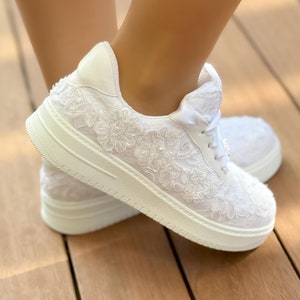 May include: White lace sneakers with a white ribbon tied around the ankle. The sneakers have a white sole and are decorated with white lace flowers.
