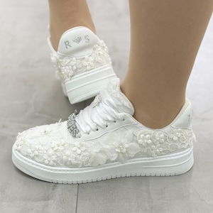 May include: White lace sneakers with pearl and flower embellishments. The sneakers have a white lace design with pearl and flower accents. The back of the shoe has the letters "R" and "S" with a heart between them.