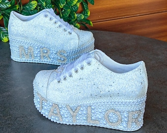 May include: White platform sneakers with a glittery finish and pearl embellishments. The words "MRS" and "TAYLOR" are spelled out in pearls on the shoes.
