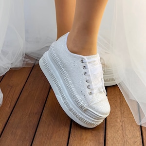 May include: White platform sneakers with a sparkly finish and pearl embellishments. The sneakers have a white satin ribbon tied around the laces.