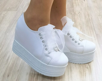May include: White platform sneakers with a white tulle bow on the laces. The sneakers have a thick platform sole and a white leather upper.