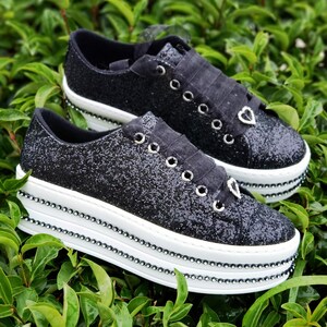 May include: A pair of black glitter platform sneakers with a white platform sole and a black ribbon tied around the ankle. The sneakers have a heart-shaped charm on each shoe.