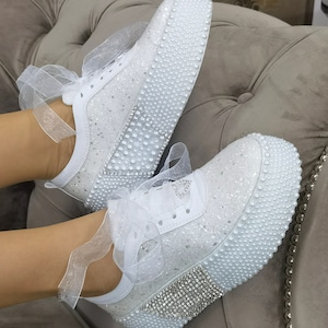May include: White sneakers with a pearl and rhinestone embellishment. The sneakers have a platform sole and a white satin ribbon.