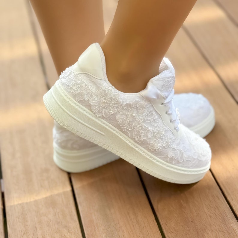 May include: White lace sneakers with a white ribbon tied around the ankle. The sneakers have a white sole and are decorated with white lace flowers.