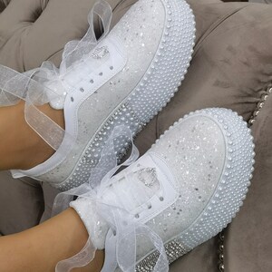 May include: White sneakers with a sparkly finish and pearl trim. The shoes have white satin ribbons tied around the ankles.