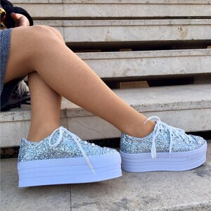 May include: Silver glitter platform sneakers with white laces and a white sole. The shoes have a chunky, layered sole design. The sneakers are a fashionable footwear choice.