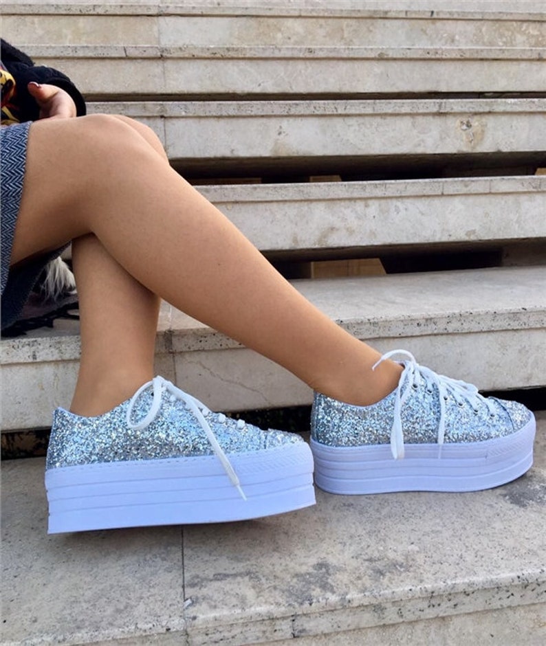 May include: Silver glitter platform sneakers with white laces and a white sole. The shoes have a chunky, layered sole design. The sneakers are a fashionable footwear choice.