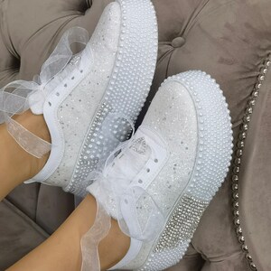 May include: White sneakers with a glittery finish and pearl embellishments. The sneakers have white laces and a white ribbon tied around the ankle.