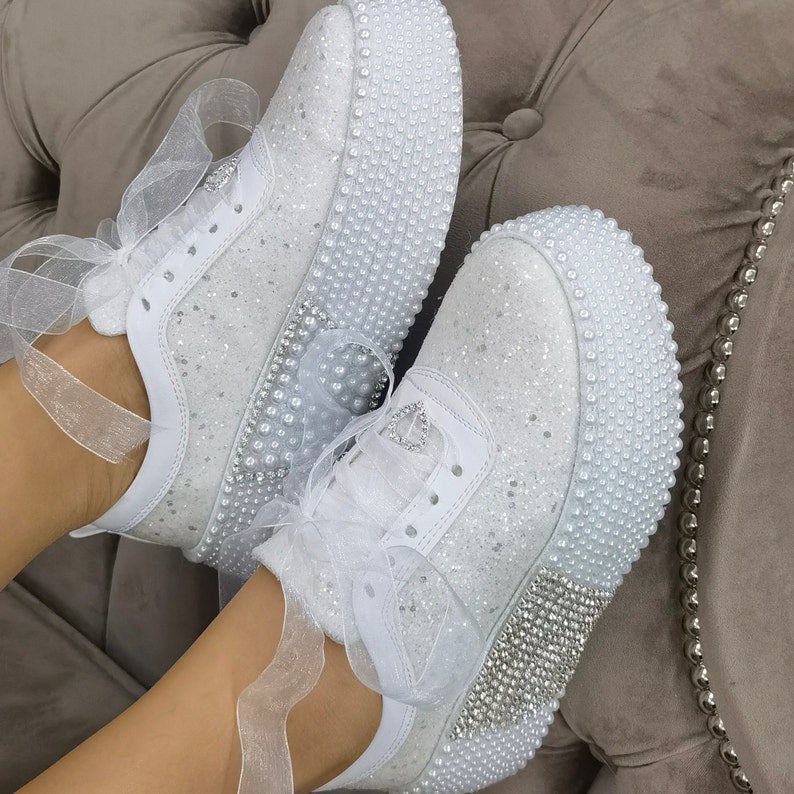 May include: White sneakers with a glittery finish and pearl embellishments. The sneakers have white laces and a white ribbon tied around the ankle.
