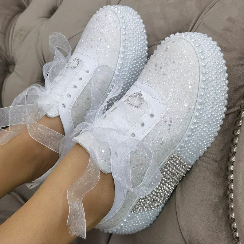 May include: White sneakers with a pearl trim and a white satin ribbon. The sneakers are covered in glitter and have a rhinestone trim.
