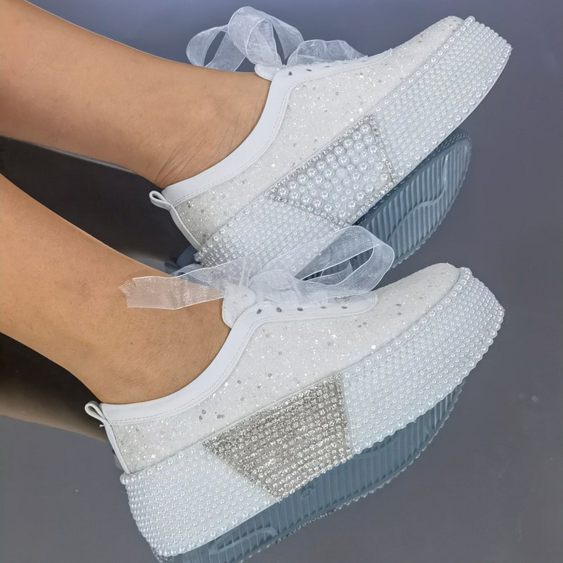 May include: White sneakers with a pearl and rhinestone embellishment. The sneakers have a platform sole and a white satin ribbon.